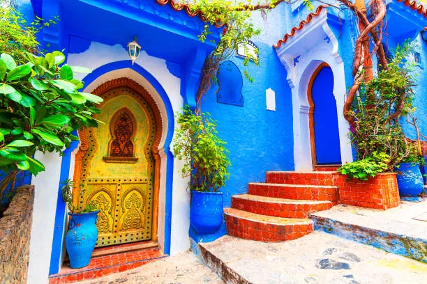 A stunning, sunlit blue alleyway in Chefchaouen Morocco, with colorful flower pots contrasting against the blue walls.
