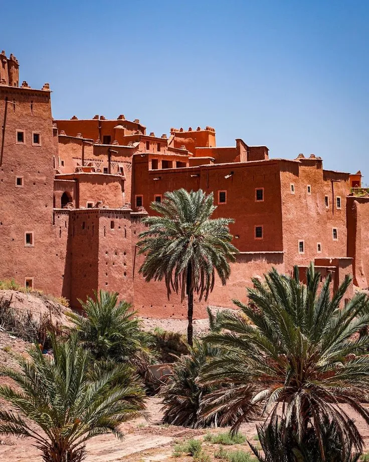 Majestic view of the red clay buildings and towers of the UNESCO World Heritage site Ait Ben Haddou framed by palm trees.