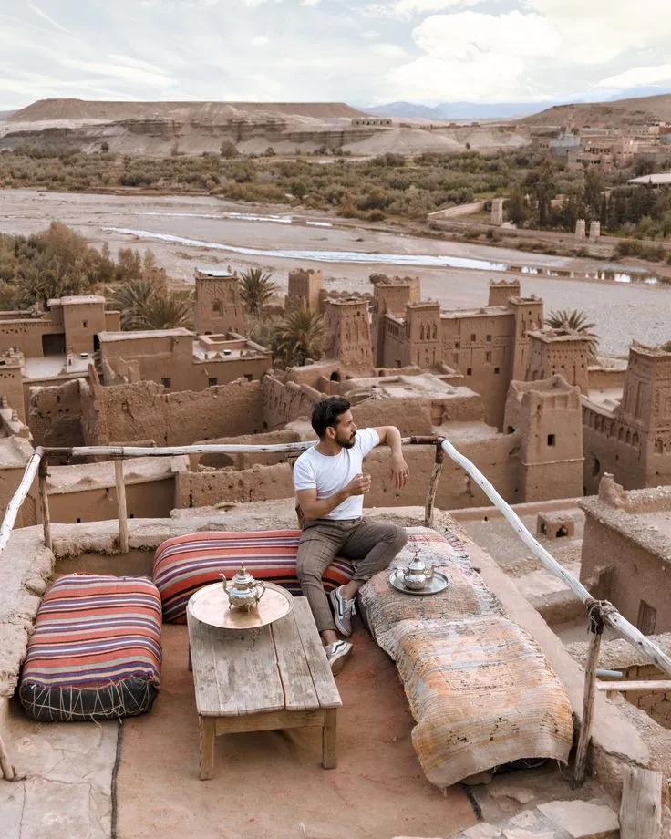 Man enjoying traditional Moroccan mint tea on a terrace with rugs and cushions, overlooking the clay architecture of the Atlas Mountains.