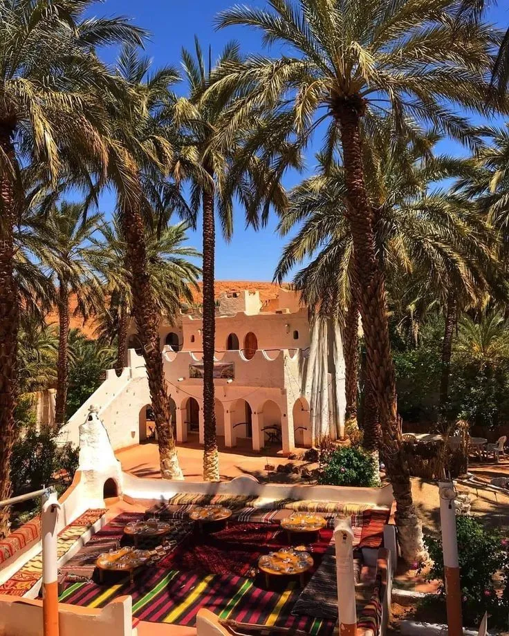 Outdoor dining area set with colorful rugs and low tables in a lush palm grove oasis outside a traditional adobe Kasbah hotel.