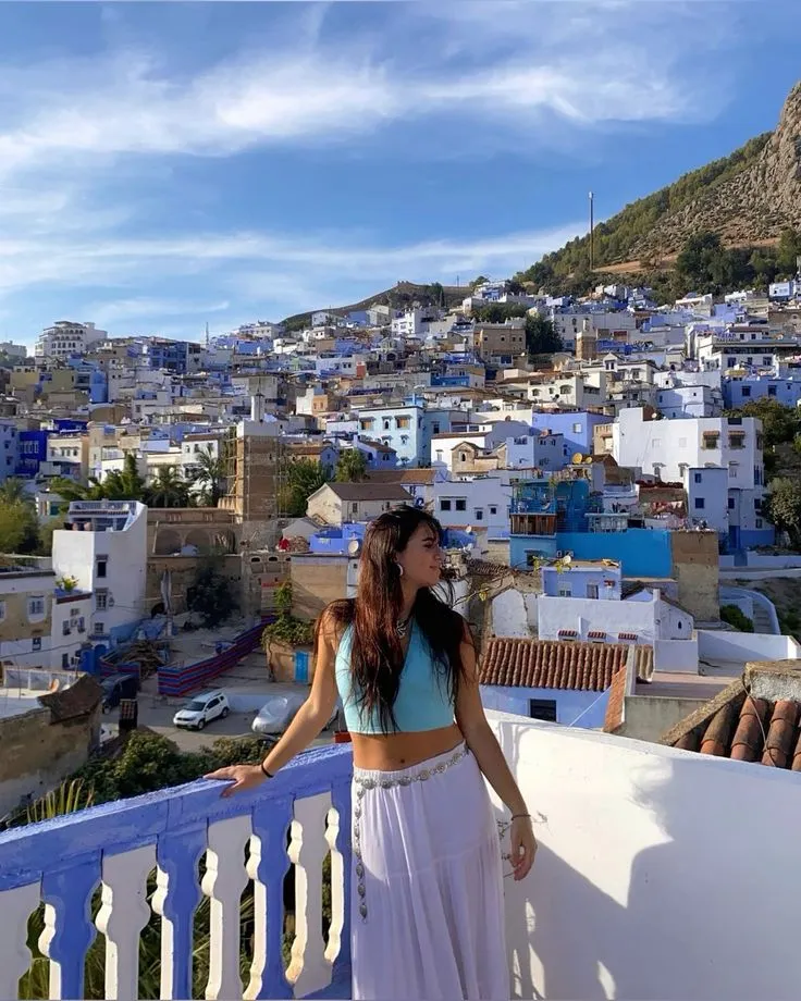 Panoramic rooftop view of the Blue City, Chefchaouen, with a traveler standing on a balcony overlooking the Rif Mountains.