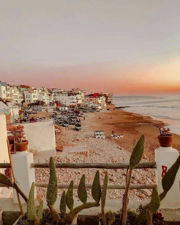 Scenic sunset over Taghazout beach featuring traditional blue fishing boats on the sand and white hillside village buildings.