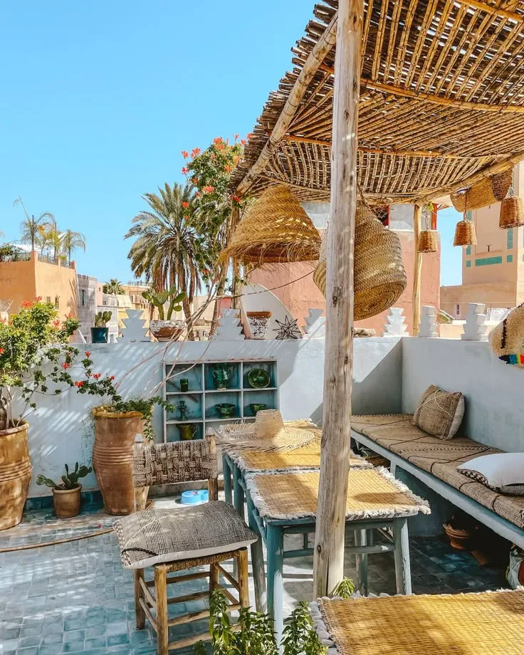 Sunny bohemian rooftop terrace in a Moroccan Riad featuring bamboo shading, wicker pendant lights, and cozy seating areas.