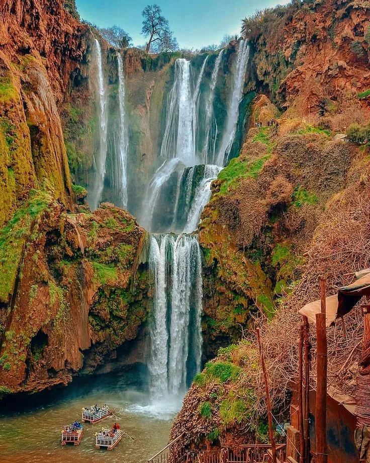 The majestic multi-tiered Cascades d'Ouzoud waterfalls flowing over red cliffs with small tourist boats navigating the pool below.