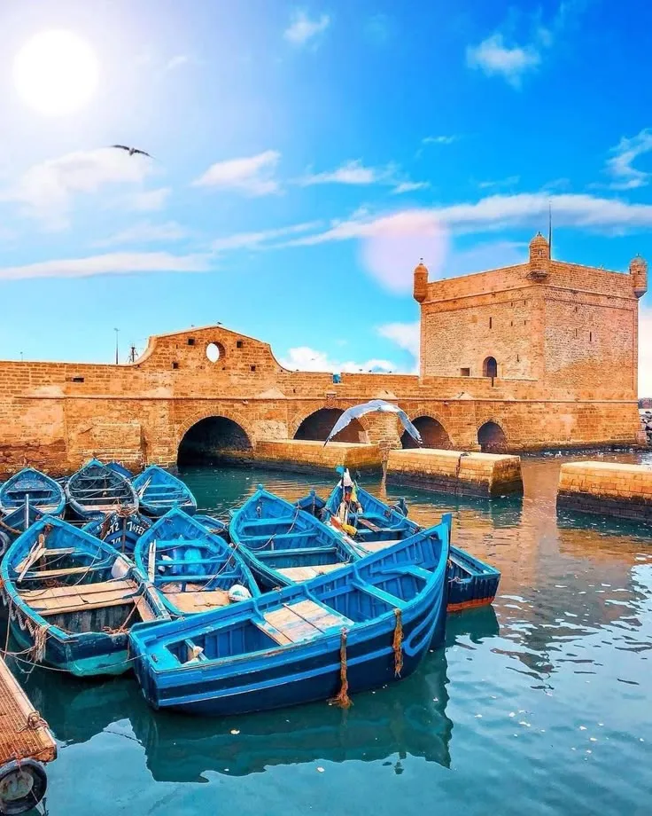 Traditional blue fishing boats docked in the harbor of Essaouira, Morocco, with the historic Skala de la Ville fortress in the background.