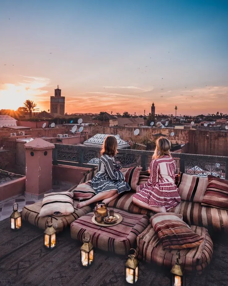 Travelers relaxing on rooftop cushions watching the sunset over the Marrakech Medina with the Koutoubia Mosque minaret in the distance.