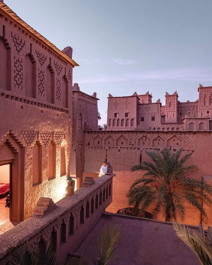 View of traditional clay architecture and geometric brickwork of a Kasbah in Southern Morocco at dusk, with a traveler on the balcony.