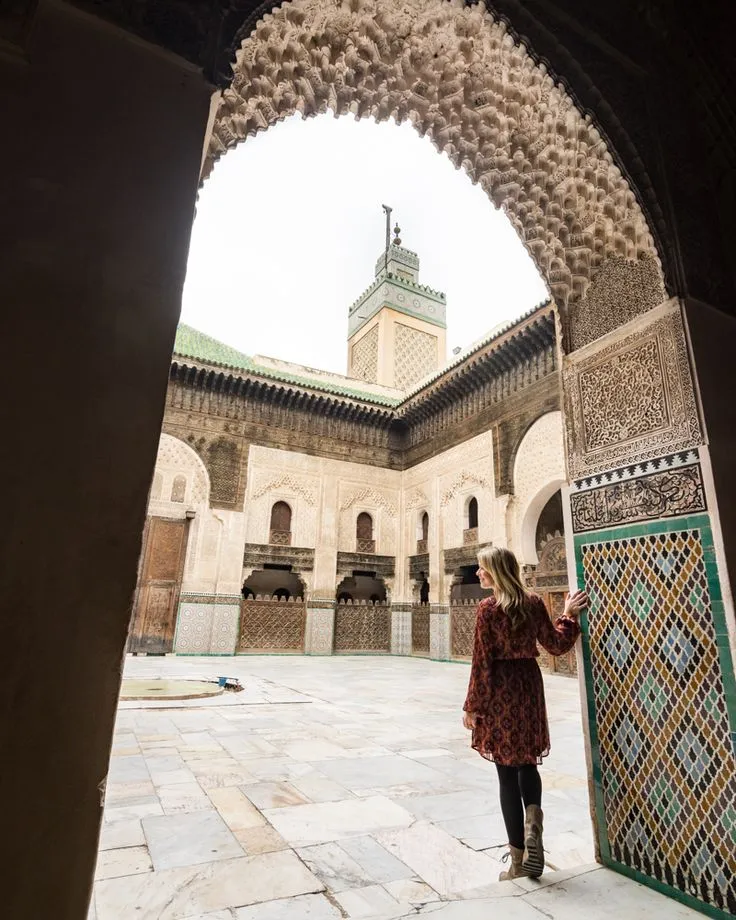 Woman admiring intricate Islamic architecture, stucco carving, and mosaic tiles inside a historic Madrasa courtyard in Fes, Morocco.