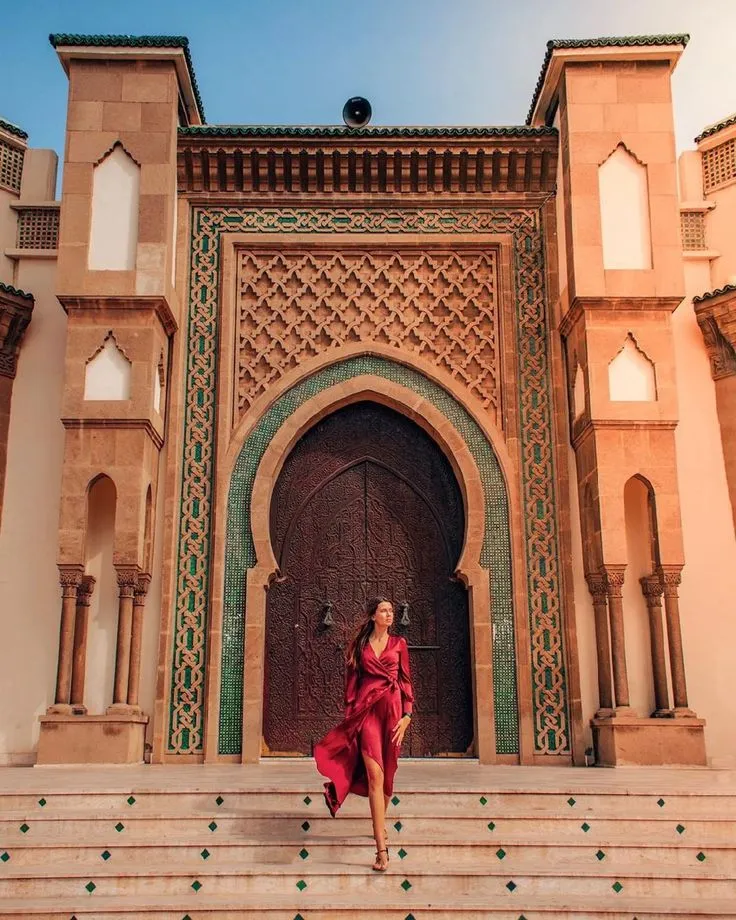 Woman in a red dress walking down the steps of the ornate Royal Palace (Dar al-Makhzen) gates in Fes, featuring detailed brass doors and Zellige tiles.