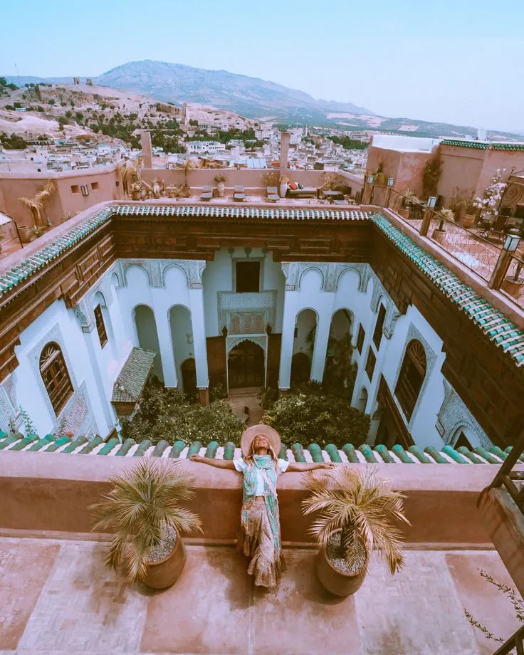 Woman in a sun hat stretching her arms on a high balcony overlooking the intricate tile work and arches of a historic Riad courtyard.