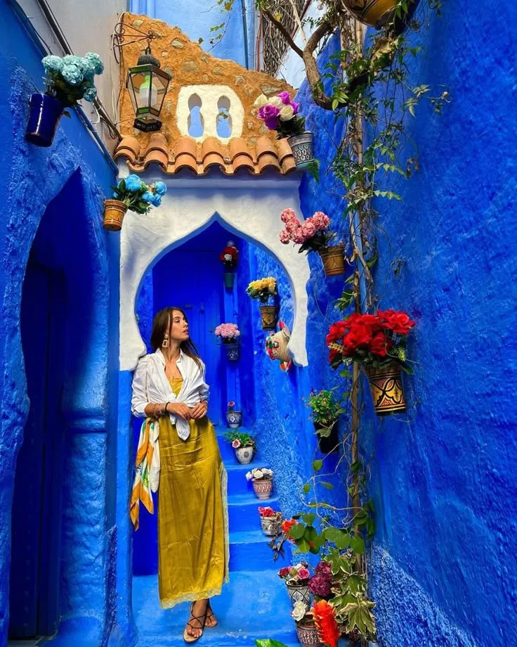 Woman in a yellow dress posing in a vibrant blue alleyway in Chefchaouen, Morocco, adorned with colorful flower pots.