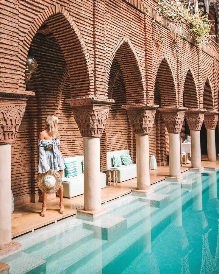 Woman standing by a luxurious indoor pool framed by traditional red brick arches and columns in a high-end Moroccan spa.