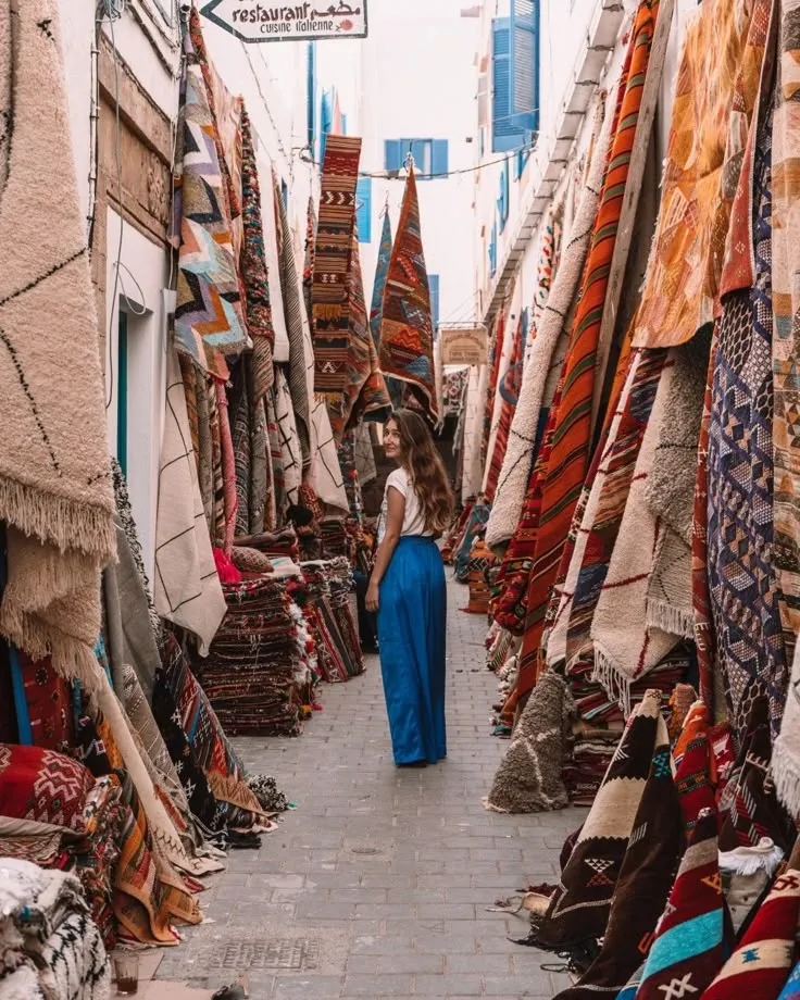 Woman standing in a narrow souk alleyway lined with colorful handmade Moroccan rugs and carpets hanging from the walls.