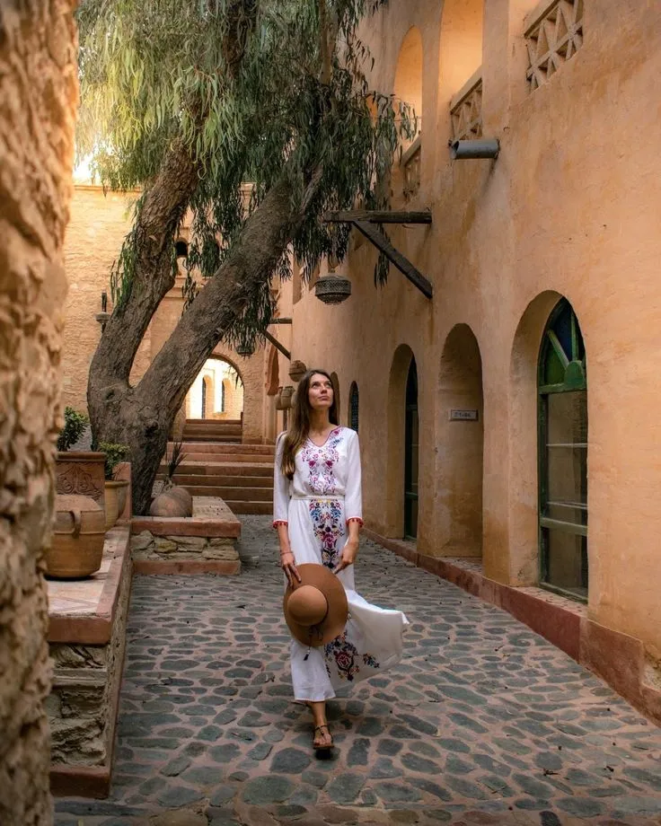 Woman walking through a quiet, earth-toned alleyway in a traditional Moroccan Medina, shaded by an old tree.