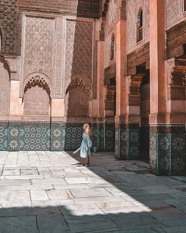 Woman walking through a sunlit corridor of a Madrasa, highlighting the Zellige tile mosaics on the lower walls.
