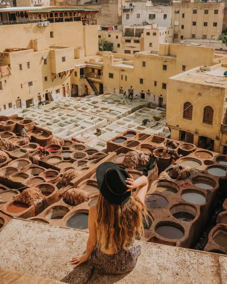 Woman wearing a black hat sitting on a terrace wall overlooking the famous honeycomb vats of the Chouara Tannery in Fes.